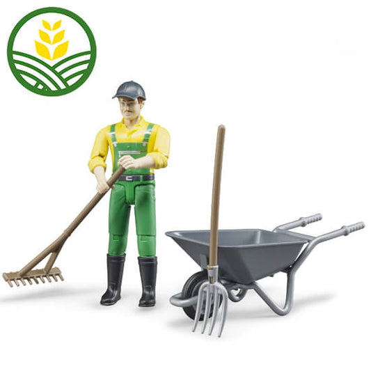 A toy male figure dressed in yellow and green John Deere overalls with a rake, pitch fork and wheelbarrow