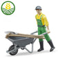 A male toy figure dressed in yellow and green overalls carrying a wheelbarrow with a rake and pitchfork resting on top.