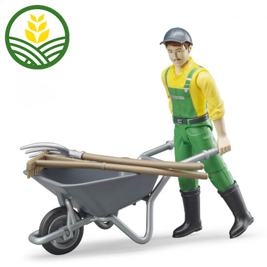 A male toy figure dressed in yellow and green overalls carrying a wheelbarrow with a rake and pitchfork resting on top.